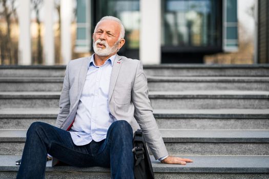 Senior businessman is tired of work. He is sitting on stairs in front of company building.