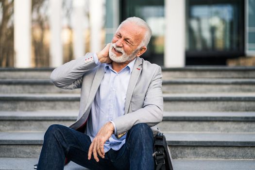 Senior businessman is tired of work. He is sitting in front of company building and having pain in neck.