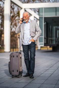 Happy senior businessman is arriving from business trip. He is talking on phone.