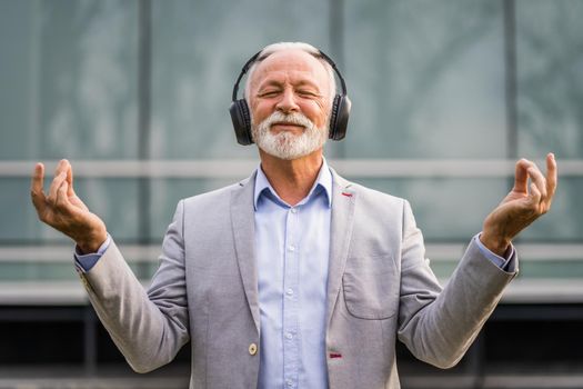 Outdoor portrait of senior businessman who is enjoying music on headphones and meditating.