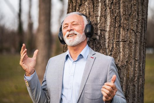 Outdoor portrait of senior businessman relaxing in park. He is listening music on headphones.