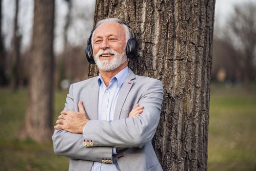 Outdoor portrait of senior businessman relaxing in park. He is listening music on headphones.