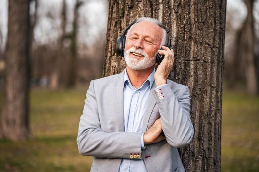 Outdoor portrait of senior businessman relaxing in park. He is listening music on headphones.