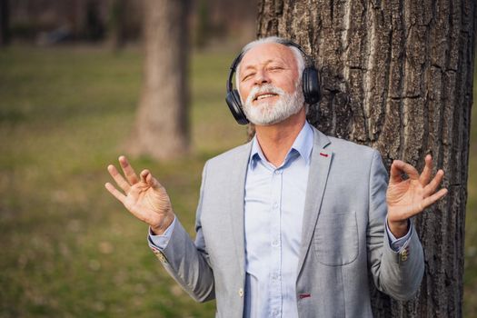 Outdoor portrait of senior businessman relaxing in park. He is listening music on headphones.