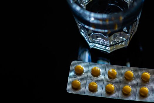 Yellow tablets in blister pack next to glass of water  on black background