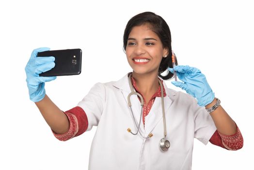 Female doctor taking selfie with stethoscope and injection by smartphone on white background.