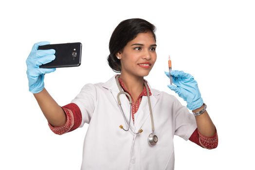 Female doctor taking selfie with stethoscope and injection by smartphone on white background.