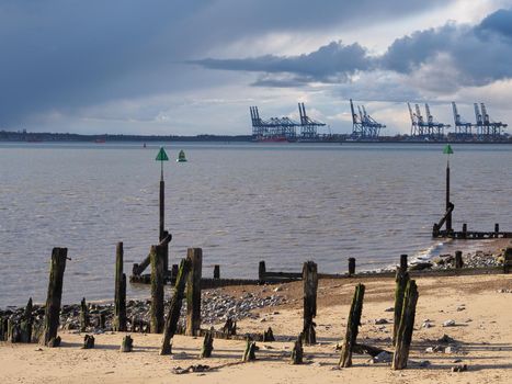 Harbour and Port of Felixstowe at dusk with remains of old wooden jetty, channel markers, groins, and cranes under dark clouds loading containers onto a cargo ship, Felixstowe, Suffolk, UK