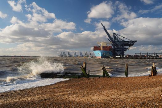 Port of Felixstowe, Suffolk, UK, March 10 2019: Cranes loading containers onto Marseille Maersk cargo ship with waves breaking over old wooden jetty