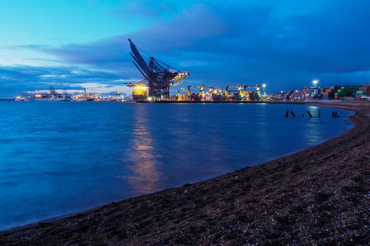 Port of Felixstowe, Suffolk, UK, October 11 2020: Cranes loading containers onto the cargo ships against the dock lights at dusk with the remains of the old wooden jetty in the foreground