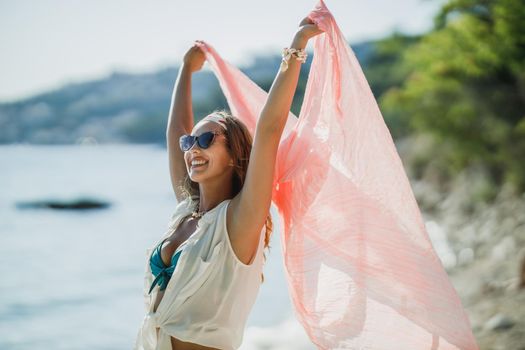 An attractive young woman is having fun and enjoying a summer vacation. She is posing with transparent scarf in her hands on the beach.