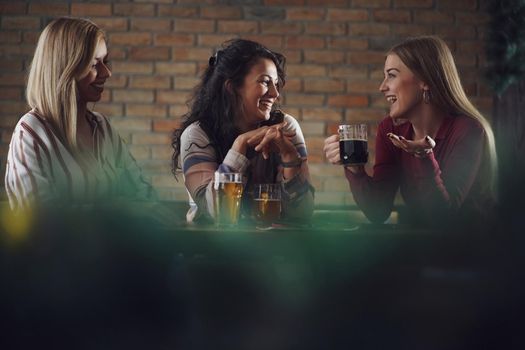 Three happy girlfriends are having fun time in pub. They are talking and drinking beer.