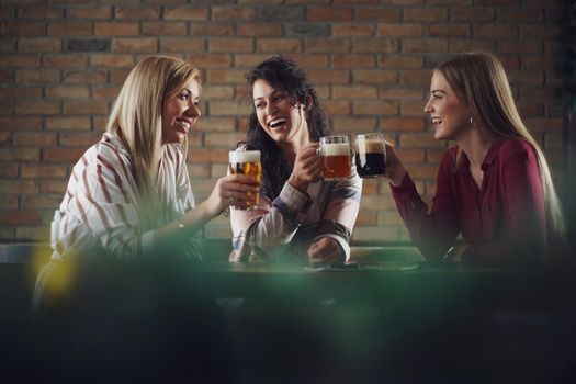 Three happy girlfriends are having fun time in pub. They are talking and drinking beer.