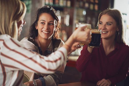 Three cheerful girlfriends are having fun time in pub. They are toasting.