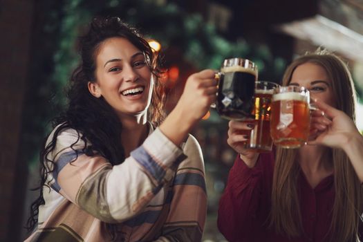 Three cheerful girlfriends are having fun time in pub. They are toasting.