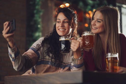 Two happy girlfriends are having fun time in pub. They are drinking beer and taking selfie. Friendship concept.