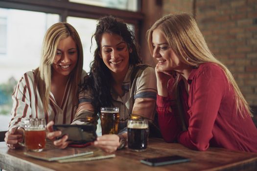 Three happy girlfriends are having fun time in pub. They are talking and drinking beer.