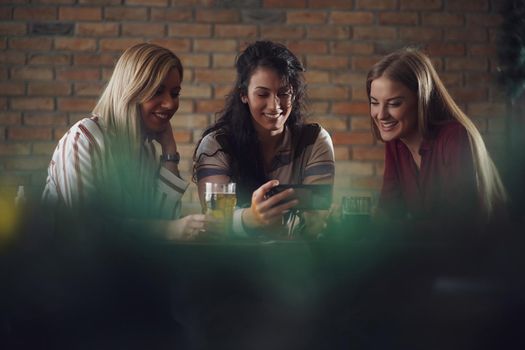 Three happy girlfriends are having fun time in pub. They are talking and drinking beer.