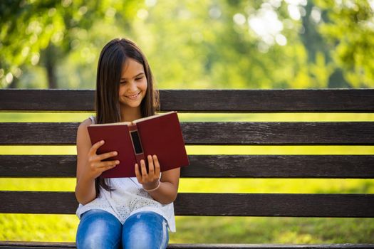 Happy little girl is reading a book in park.