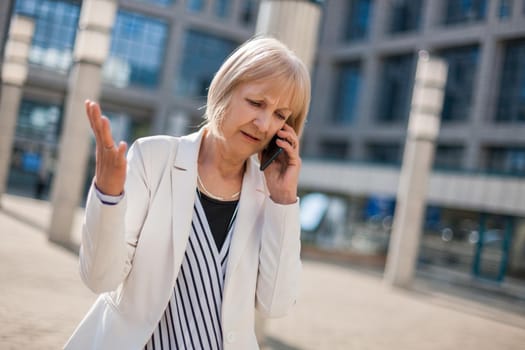 Outdoor portrait of angry senior businesswoman who is talking on phone.