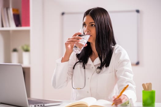 Young doctor is working in her office. She is drinking water.