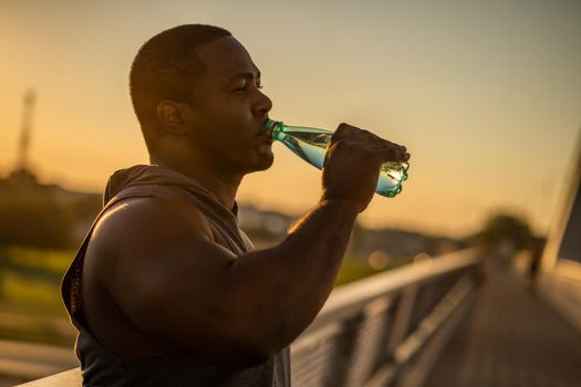 Young african-american man is drinking water after exercising on the bridge in the city.