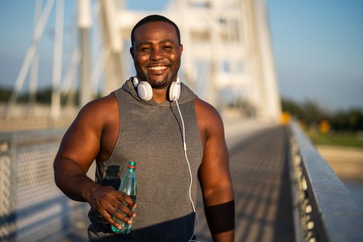 Portrait of young cheerful african-american man in sports clothing who is drinking water after exercising.