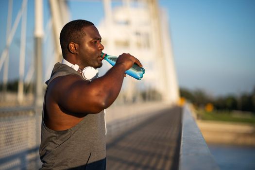 Portrait of young cheerful african-american man in sports clothing who is drinking water after exercising.