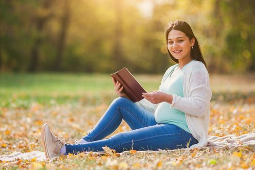 Pregnant woman relaxing in park. She is reading book.