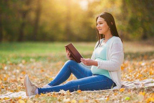 Pregnant woman relaxing in park. She is reading book.