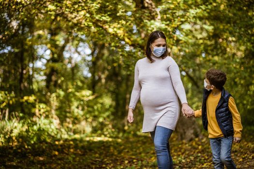 Pregnant woman and her son are walking in park in autumn. They are wearing protective face mask. Covid-19 pandemic concept.