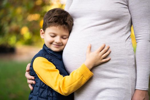 Boy is leaning on stomach of his pregnant mom in park in autumn. Family relaxing time in nature.