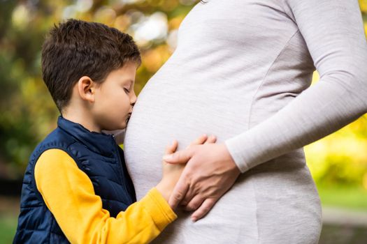 Boy is kissing stomach of his pregnant mom in park in autumn. Family relaxing time in nature.