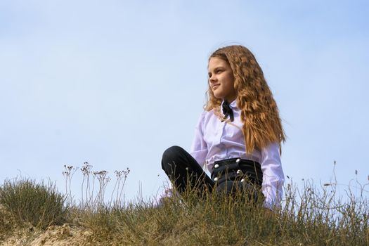A girl in a white shirt with a bow tie sits on the ground in a field against the background of the sky