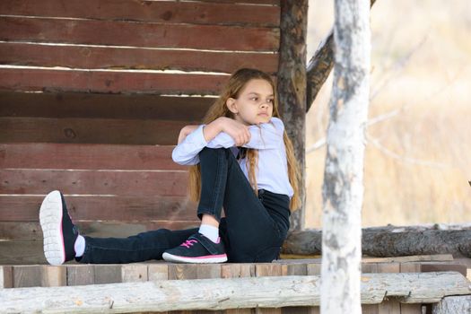 A girl sits on a wooden table against the background of a wall of boards and looks into the distance