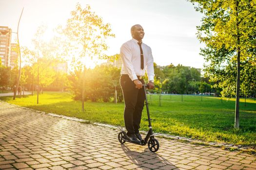 Happy african-american businessman is riding scooter in park on sunny day.