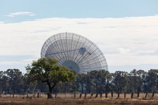 A large outdoor scientific radio telescope in the sunshine in a green field with trees