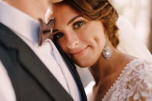 A close-up portrait of the bride looking at the camera, put her head on the breast of the groom. A beautiful wedding couple.