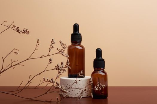 Two dark brown bottles with skin serums to cure acne and moisturize skin. Cosmetic bottles on beige stone podium and dark brown table against beige background decorated with dry flowers. 