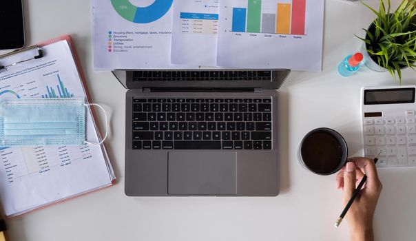 Top view of male hands holding a cup of coffee working on laptop calculator financial report and medical face mask, business accounting with new normal during Covid 19 pandemic concept