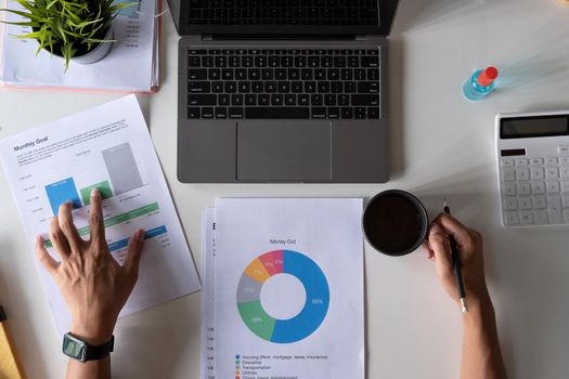 Top view of male hands holding a cup of coffee working on laptop calculator financial report and medical face mask on desk, business accounting with new normal during Covid 19 pandemic concept.