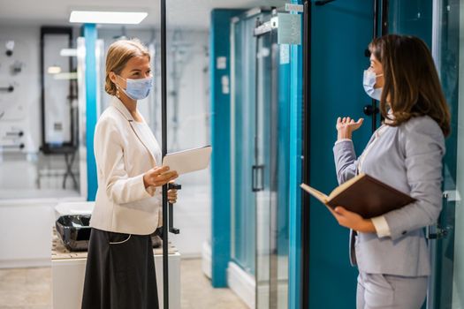 Businesswoman owning small business bath store. She is talking with a customer who is choosing the goods. They are wearing protective face mask.