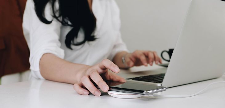 Cropped image of an office woman is charging a smartphone with a wireless charger while typing on a computer laptop..