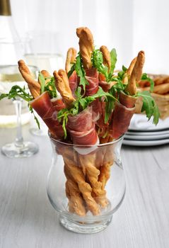 Grissini - breadsticks with Parmesan, wrapped with a piece of prosciutto and arugula. Italian dish with antipasto on table.