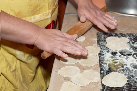Hands knead the dough on the kitchen table with a wooden rolling pin. Roll out dough for making dumplings