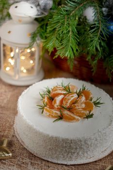 A large round cake decorated with tangerine slices on a New Year table with a flashlight and a spruce branch. Vertical frame