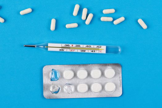 Mercury glass thermometer, white capsules and tablets in packaging on a blue background. View from above