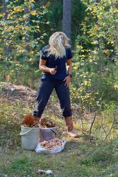 A blonde woman with a knife in her hand and her hair loose is standing in the forest next to the collected mushrooms.