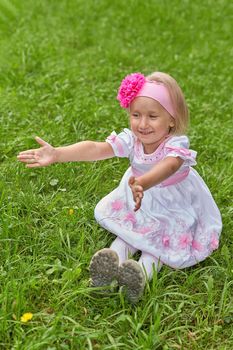 Portrait of a little girl in a beautiful dress and a bow. The child sits on the grass and stretches out his arms forward.