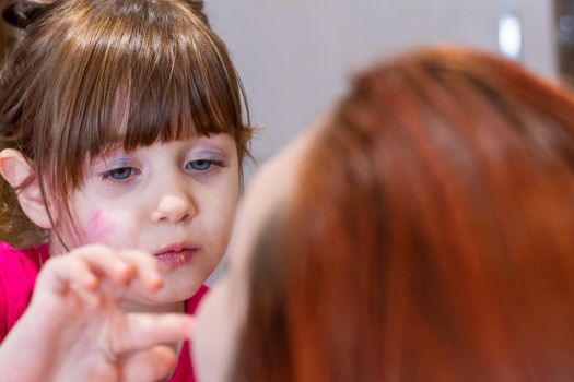 Protrait of a cute, blue-eyed, brown-haired girl with makeup in her face playing to put makeup in a redhead woman